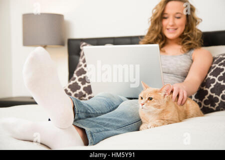 Teenage girl (16-17) lying on bed with laptop and stroking ginger tabby cat Stock Photo