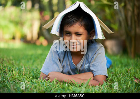 small smiling boy with book on head lay on grass Stock Photo