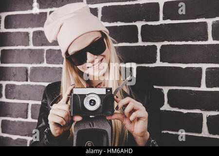 Portrait of pretty blond hair smileing young teenage hipster woman model with retro photo camera wearing a pink hat, black leather jacket. Fashion look. Stock Photo