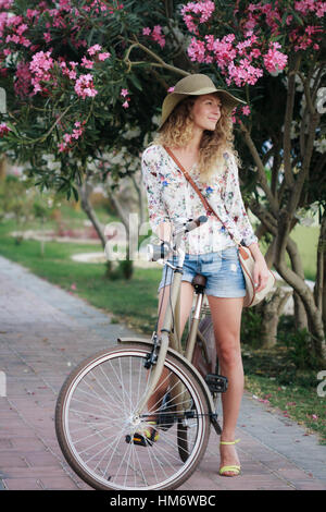 Blonde girl wearing hat and shorts with vintage bicycle and pink oleander tree behind her in park. Happy girl on vacation in resort town Stock Photo