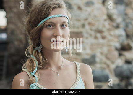 Close up portrait of beautiful blonde girl with blue eyes and braid wearing blue earrings and turquoise headband against blurred background of stone w Stock Photo