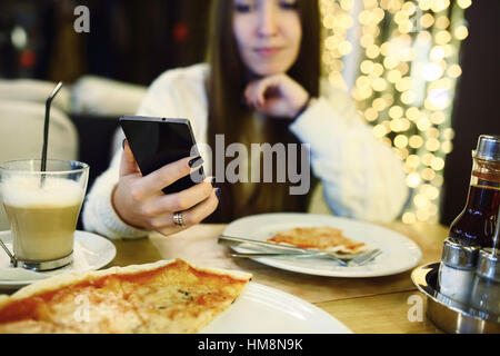 Woman typing write message on smart phone in a modern cafe. Cropped image of young pretty girl sitting at a table with pizza using mobile phone. Tonned. Selective focus. Stock Photo
