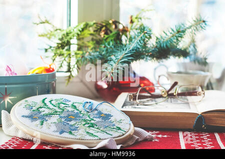 Winter still life : hoop with embroidered flowers pattern, open book, glasses, teapot and cup, colorful yarn in box and christmas tree in vase. Select Stock Photo