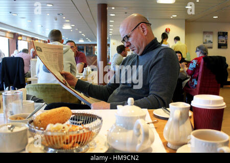 Middle aged man reading newspaper whilst having breakfast on board a ferry. Stock Photo