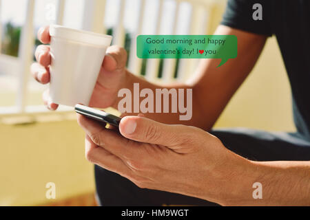 closeup of a young caucasian man with a cup of coffee in his hand sending or reading a text message with a smartphone with the text good morning and h Stock Photo