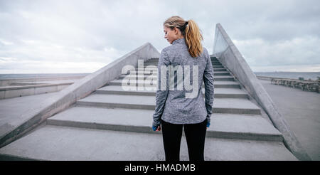 Rear view shot of young female standing by a stairs before workout. Fitness female model standing by steps in morning. Stock Photo