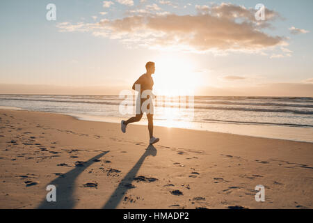 Image of young runner running in morning along the beach. Young man jogging on the sea shore at sunrise. Stock Photo