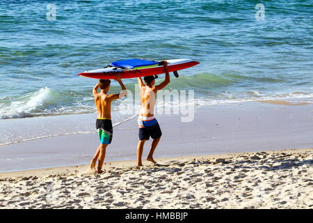 Two young boys carry a red surf rescue board on their heads along the sand at Kings Beach, Caloundra on the Sunshine Coast of Queensland, Australia. Stock Photo