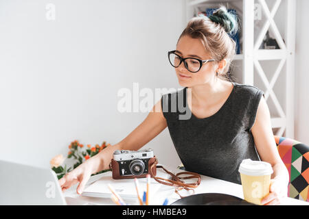 Attractive young woman photogtrapher in glasses drinking coffee and using laptop at the table in the room Stock Photo