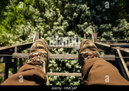 View looking down from the top of an abandoned railroad trestle roughly 80 feet off the ground above trees and brush below. Focused on feet in brown a Stock Photo