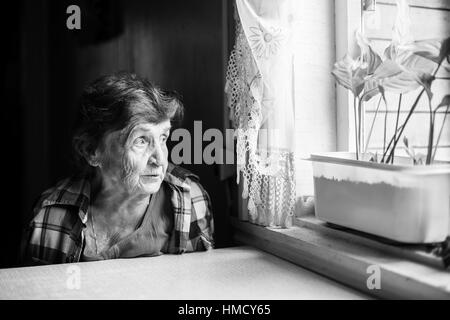 An elderly woman sadly looking out the window. Black-and-white photo. Stock Photo