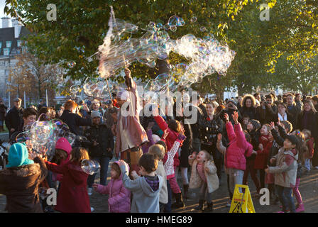 Small children and their parents gather round a street performer leaving a trail of giant bubbles on the South bank in London Stock Photo