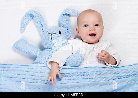 Funny little baby wearing a warm knitted jacket playing with toy bunny relaxing on white cable knit blanket in sunny nursery Stock Photo