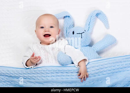 Funny little baby wearing a warm knitted jacket playing with toy bunny relaxing on white cable knit blanket in sunny nursery Stock Photo