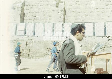 View facing north of a male Israeli soldier, wearing a kippah (yarmulke), tallit prayer shawl, and tefillin wrapped on his arm, praying while facing the Western Wall, in Jerusalem, Israel, November, 1967. Israeli school boys wearing traditional blue tembel hats are walking in the background toward the Wall. (Photo by Morse Collection/Gado/Getty Images). Stock Photo