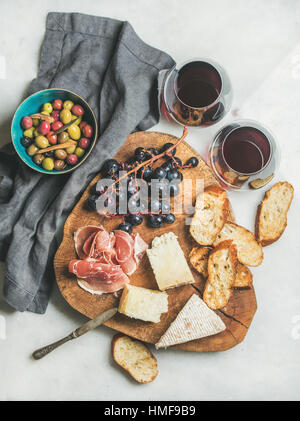 Wine and snack set. Variety of cheese, olives in blue bowl, prosciutto, roasted baguette slices, black grapes on wooden board and glasses of red wine  Stock Photo