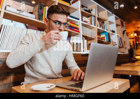 Image of young attractive man sitting in cafe while drinking coffee and using laptop. Stock Photo