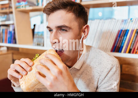 Image of young hungry man dressed in white shirt sitting in cafe while eating sandwich and listening music. Stock Photo
