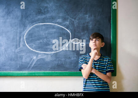 Concepts on blackboard at school. Intelligent and smart hispanic boy in class. Portrait of male kid thinking with hand on chin, looking up against clo Stock Photo