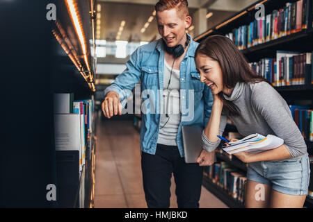 Image of happy young man and woman standing by book shelf in library and looking for books. Students at college library looking for books.
