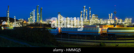 Panoramic view of a chemical plant and refinery with night blue sky and illumination, some freight trains in the foreground. Stock Photo