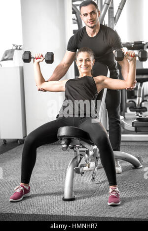 Young adult fitness couple. Woman doing chest and shoulder dumbbell  press on bench with personal coach assisting her and giving instructions. Stock Photo