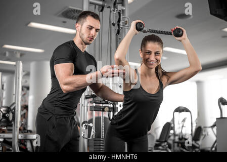 Young adult personal fitness trainer supporting his client while doing overhead rope extension. Stock Photo