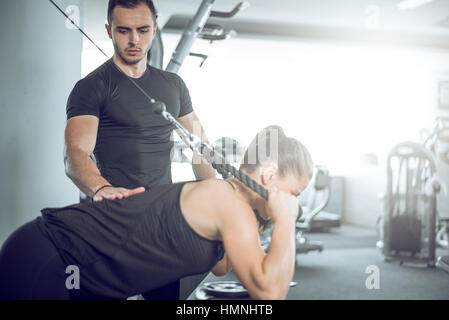 Young adult personal fitness trainer supporting his client while doing overhead rope extension. Stock Photo