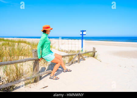 Young woman sitting on wooden fence and looking at beautiful white sand beach, Baltic Sea, Poland Stock Photo