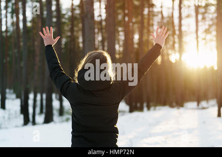 teen girl raised hands from behind in winter pine forest in sunset Stock Photo