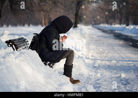 lonely person with hoodie and backpack staying on a bench in city park alley with snow in winter time and being cold Stock Photo