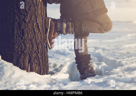 horizontal close up of woman wearing brown boots, textile gaiters and blue jeans leaning against a large tree covered in snow in winter time, concept  Stock Photo