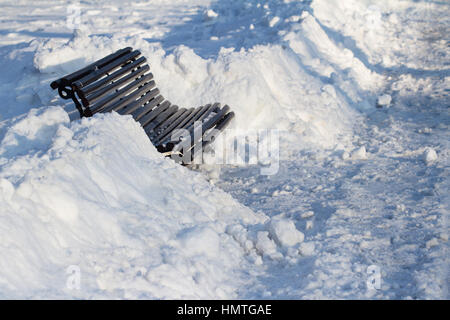 perspective view of wooden black bench in a city park alley covered in snow, winter time Stock Photo