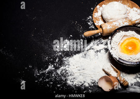 Ingredients and utensils for the preparation of bakery products - flour, dough, eggs, rolling pin, whisk, strainer, bread - on black table with free c Stock Photo