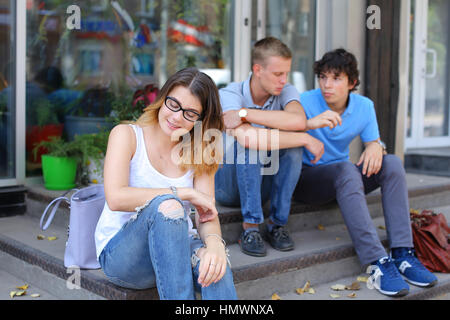 Three friend female male sitting on the doorstep floor cafe, chatting, talking, using technology, tablet, phone sun weather summer Stock Photo