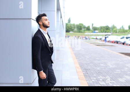 Young handsome businessman Arab Muslims is in the business center in black elegant dress on building background. Businessman man male looking at time  Stock Photo