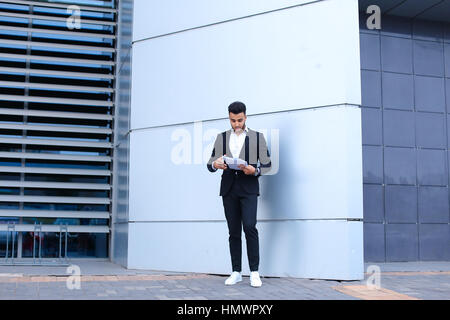 Young tall handsome arab entrepreneur guy student holds and considers, reads paper or documents, stands in full growth directly into camera near gray  Stock Photo