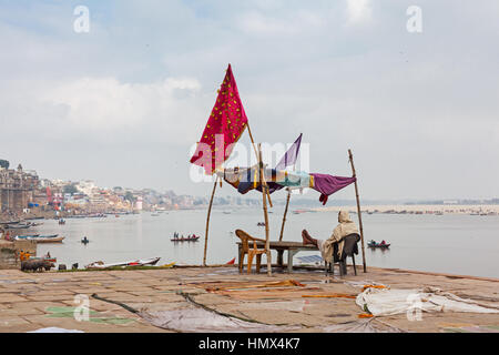 VARANASI, INDIA - JANUARY 04, 2015 : A man sits relaxing under a makeshift shelter above Narad Ghat on the Ganges river. Many small, square kites can  Stock Photo