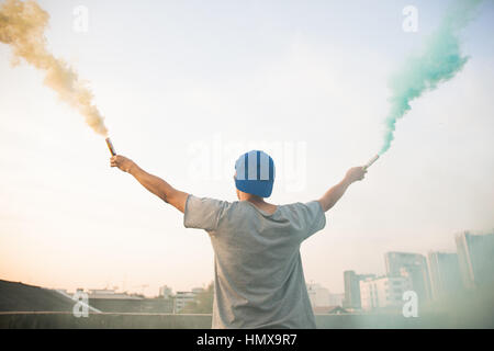 Male teenager holding colorful smoke sticks up in the air over urban city background Stock Photo