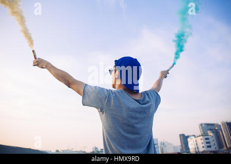 Male teenager holding colorful smoke sticks up in the air over urban city background Stock Photo
