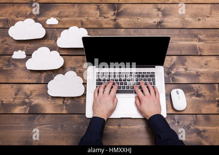 Cloud computing technology concept with white fluffy clouds next to the laptop. Mans hands typing the the keyboard uploading data, on a wooden table. Stock Photo
