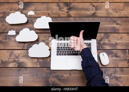 Cloud computing technology concept with white fluffy clouds next to the laptop. Man with thumbs up over the  keyboard uploading data on a wooden table Stock Photo