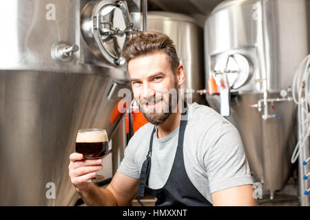 Handsome brewer in uniform tasting beer at the brewery with metal containers on the background Stock Photo