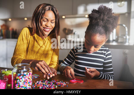 Attractive young African American female parent helping cute child in striped shirt choose bead colors for heart shape frame on table in kitchen Stock Photo