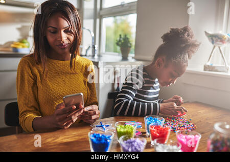 Busy African American parent in yellow sweater checking her phone while child creates beading crafts on wooden table in kitchen Stock Photo