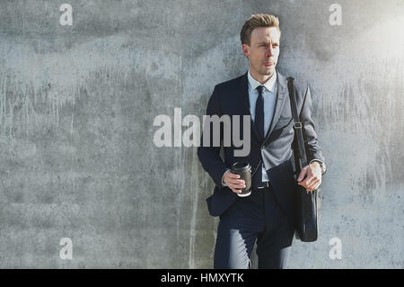 Young male in suit and handbag standing near wall with cup of coffee and looking away Stock Photo