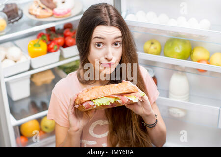 Young woman with big sandwich in front of the refrigerator full of friuts and vegetables Stock Photo