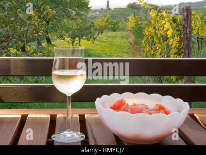 Glass of white wine and plate of fresh tomato salad on wooden table with picturesque mediterranean view in background. Brda region, Western Slovenia. Stock Photo