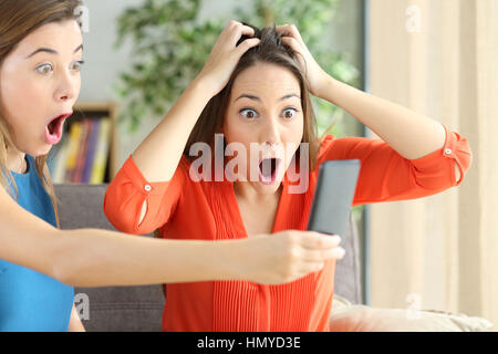 Two casual girls amazing watching on line promotions in a cellular sitting on a couch in the living room with a homey background Stock Photo