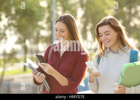 Two happy students walking together and using everyone her smart phone on line outdoors in a park or university campus Stock Photo